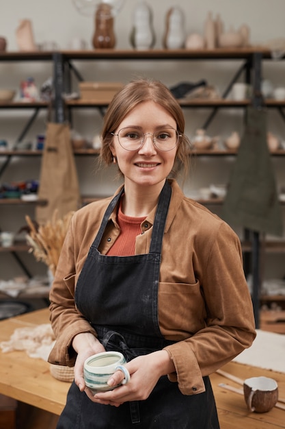 Vertical waist up portrait of young female potter looking at camera while posing in workshop, hobby and small business