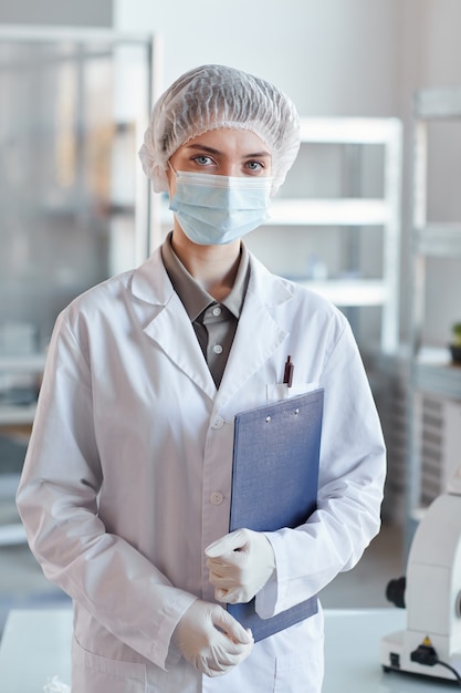 Vertical waist up portrait of young female scientist wearing face mask and looking at camera while standing in medical laboratory and holding clipboard