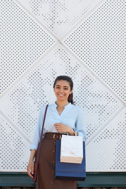 Вертикальная талия вверх портрет молодой женщины, держащей хозяйственные сумки и улыбающейся в камеру, стоя на открытом воздухе у торгового центра, скопируйте пространство выше