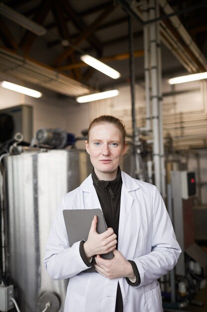 Vertical waist up portrait of young woman wearing lab coat posing against machines while working at dairy factory