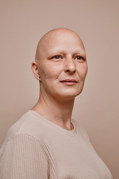 Verticale dai toni caldi ritratto di fiduciosa donna calva che guarda lontano sperando mentre posa su sfondo beige minimo in studio, alopecia e consapevolezza del cancro
