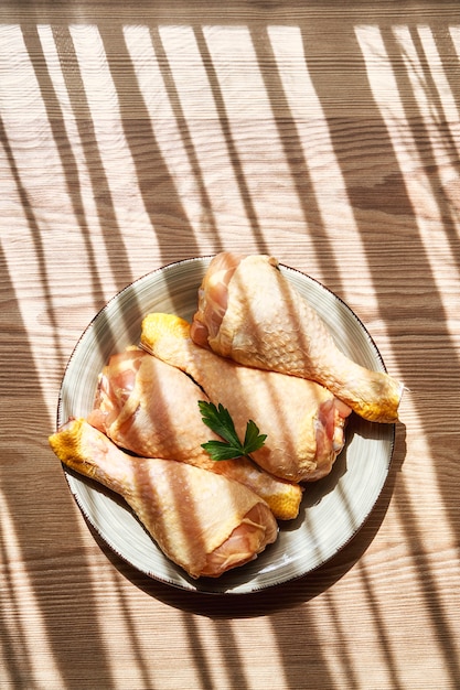 Verticale compositie van het bovenaanzicht van een bord met rauwe kippenpoten op een houten tafel naast een raam dat de zonnestralen tussen licht en schaduw doorlaat