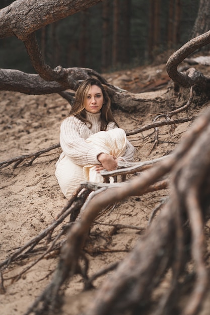 Verticale foto van een meisje in de natuur, een brunette in een lichte trui zit tussen de wortels van bomen die...