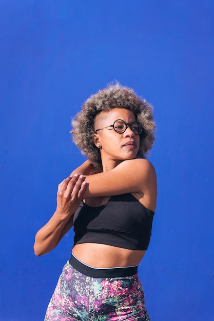 verticale foto van een serieuze Afro-Amerikaanse sportvrouw die zich uitstrekt in een blauw achtergrondconcept van sport en actieve levensstijl, kopieerruimte voor tekst