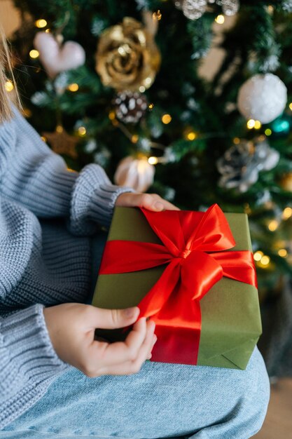 Verticale gekapte opname van een onherkenbaar klein meisje met een kerstcadeau doos terwijl het in de ochtend onder een kerstboom zit, verlicht met een krans.