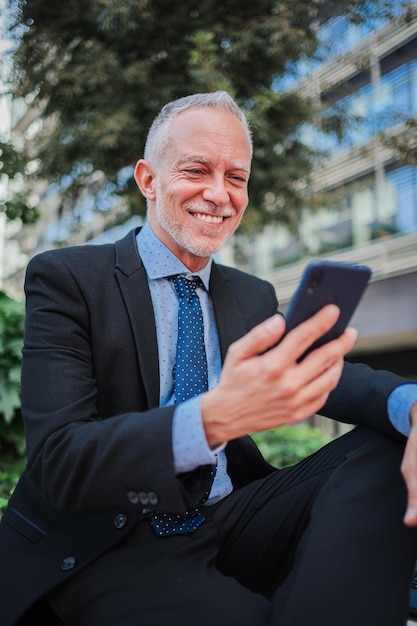 Verticale glimlachende volwassen zakenman in pak met een smartphone die buiten zit te ontspannen en