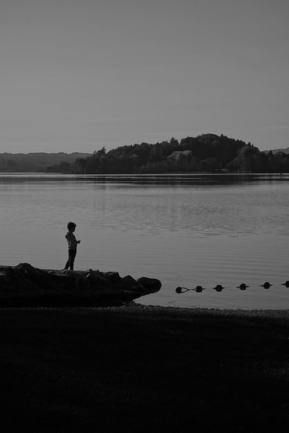 Verticale grijswaardenopname van een jongen die aan de oever van een meer staat