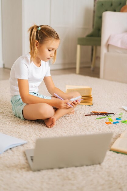 Verticale opname van een gefocust, vrij primair meisjeskind dat huiswerk schrijft in een schrijfboek dat thuis op de vloer zit