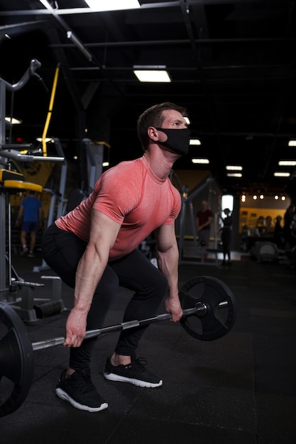 Verticale opname van een gespierde sportman met een medisch gezichtsmasker die een zware halter opheft in de sportschool