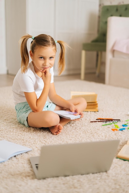 Verticale opname van een glimlachend blond, vrij primair meisje dat huiswerk schrijft in een beurt die thuis op de vloer zit en naar de camera kijkt. Schattig elementair schoolmeisje huiswerk met behulp van laptopcomputer.