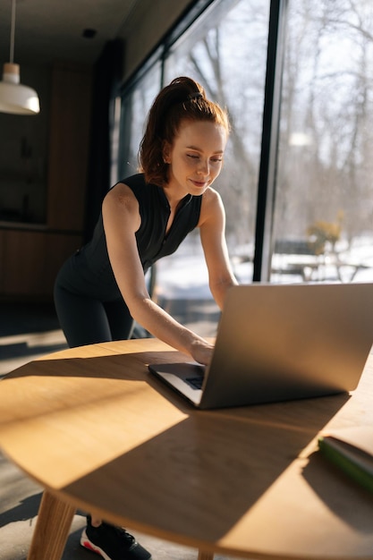 Verticale opname van een sportieve vrouw in een outfit met behulp van een laptop die op een zonnige dag bij het bureau bij het raam staat