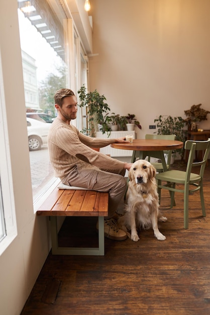 Verticale opname van jonge man eigenaar van golden retriever zit in een café met en streelt zijn hond onder