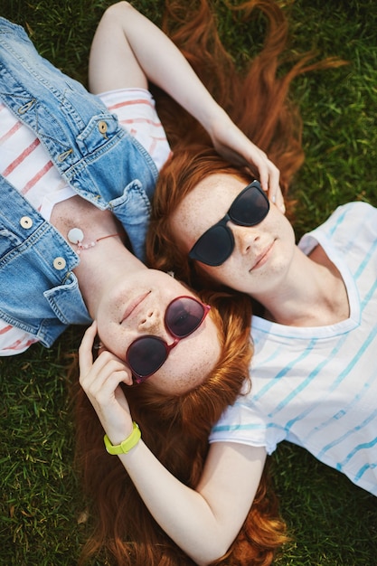 Verticale opname van twee mooie zussen met rood haar en sproeten, liggend op gras en lachend met ontspannen uitdrukking, gezichten aanraken, zorg voor elkaar uitdrukken.
