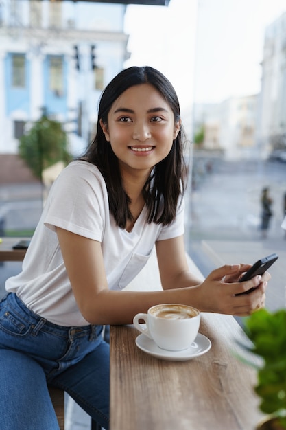 Verticale portret van aziatische vrouw zitten in stadscafé in de buurt van venster op zomerdag, mobiele telefoon vasthouden en koffie drinken, glimlachend opzij