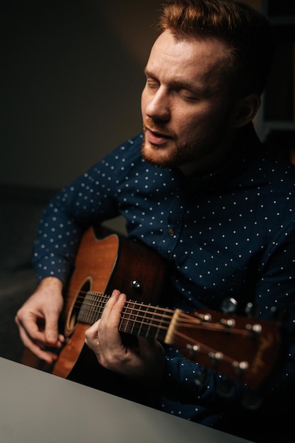 Verticale portret van gitarist zanger mannelijke akoestische gitaar spelen en zingen lied in donkere woonkamer thuis studio creatieve muzikant met vrijetijdsbesteding alleen in appartement