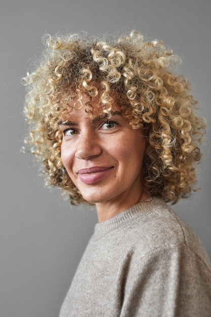 Verticale portret van glimlachende vrouw met krullend haar terwijl poseren tegen een grijze achtergrond, uniek schoonheidsconcept