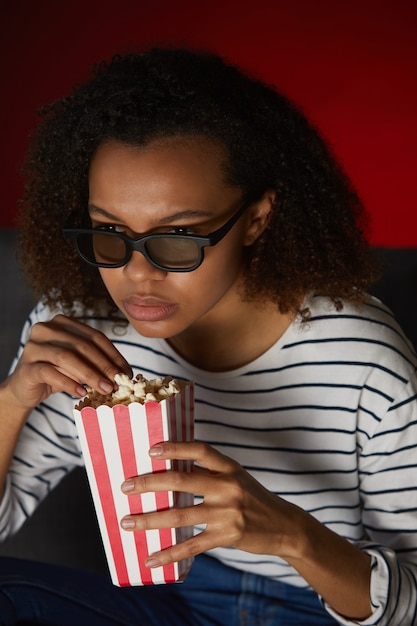 Verticale portret van jonge Afro-Amerikaanse vrouw thuis Tv kijken en popcorn eten zittend in een donkere kamer