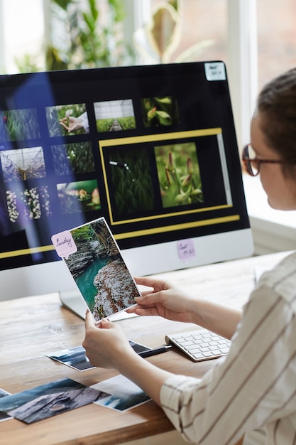 Verticale portret van jonge vrouwelijke fotograaf foto's houden en glimlachen zittend aan een bureau met fotobewerkingssoftware op computerscherm