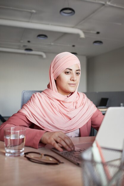 Verticale portret van jonge zakenvrouw hoofddoek dragen laptop aan balie tijdens het werken in office alleen, kopieer ruimte