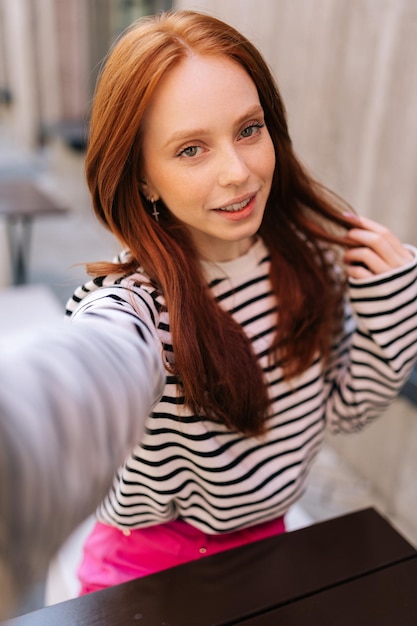 Verticale pov-portret van een mooie roodharige vrouw die een selfie maakt met de camera van een smartphone, zittend aan tafel in een café in de buitenlucht, uit het oogpunt van een charmante jonge vrouw die een selfie maakt met de camera van een mobiele telefoon