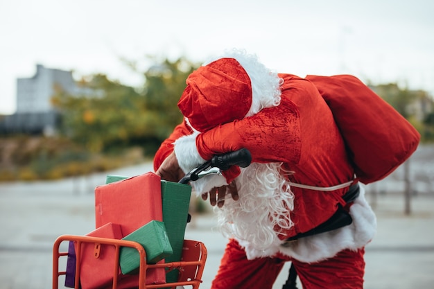 Foto verticale stockfoto van de kerstman ondersteund in een fiets vol geschenken met uitgeputte uitdrukking.