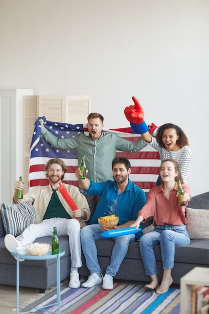 Verticale volledige lengte portret van een multi-etnische groep vrienden kijken naar sportwedstrijd op tv en emotioneel juichen terwijl ze de Amerikaanse vlag vasthouden