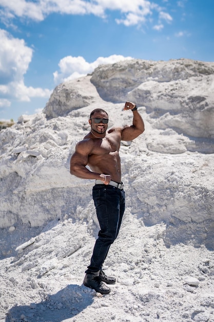 Verticale weergave van de sterk passende bodybuilder die zijn biceps laat zien. Knappe atletische man die zijn spieren laat zien terwijl hij op het zand staat met blauwe lucht op de achtergrond