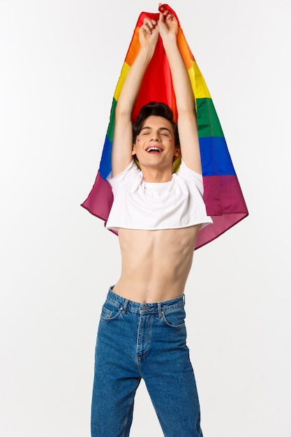 Foto verticale weergave van trotse en gelukkige homoseksuele man verhogen lgbtq regenboogvlag, glimlachend met opgelucht emotie