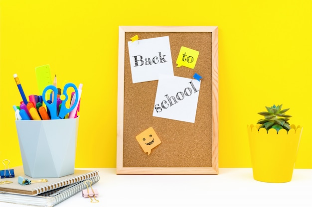 Vertically board for notes and the words Back to school on attached sticky sheets, office supplies and plant on table in classroom.