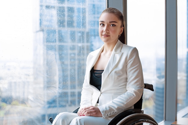 Vertrouwen gelukkig zakenvrouw in rolstoel tegen de achtergrond van een panoramisch raam met uitzicht op de wolkenkrabbers en een grote stad, ze lacht naar de camera, handicap overwinnen concept