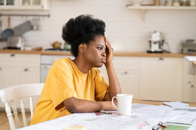 Verveeld Afrikaans meisje moe van freelance werk op afstand, zit gefrustreerd aan een thuisbureau met papieren