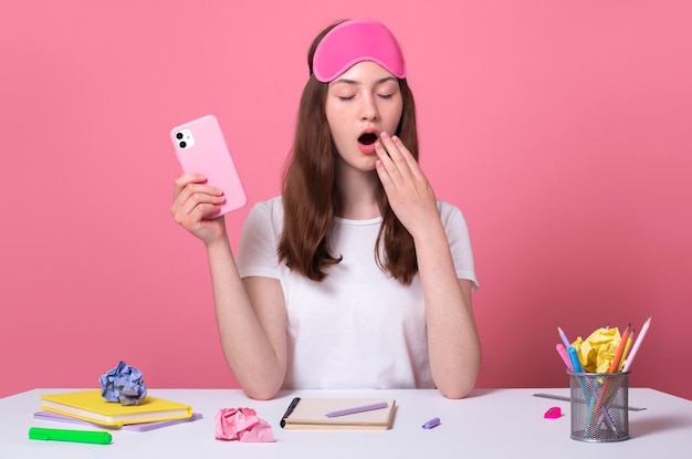 Foto verveeld geeuwen slaperig schoolmeisje dat huiswerk doet, moe van het studeren, houdt een smartphone in de hand