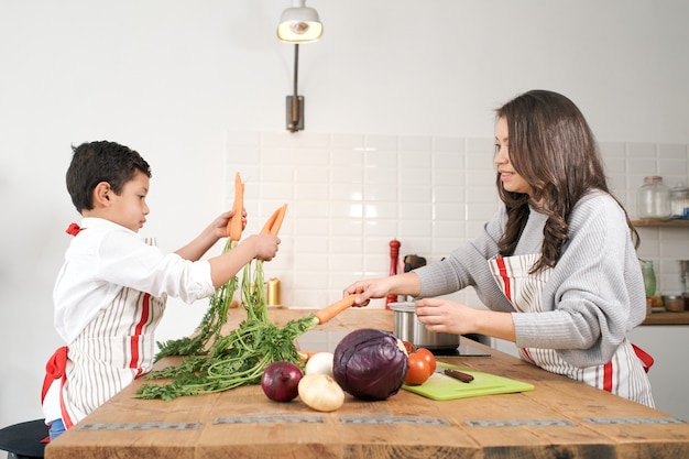 Verveeld kind speelt met wat groenten in de keuken terwijl moeder gezond kookt