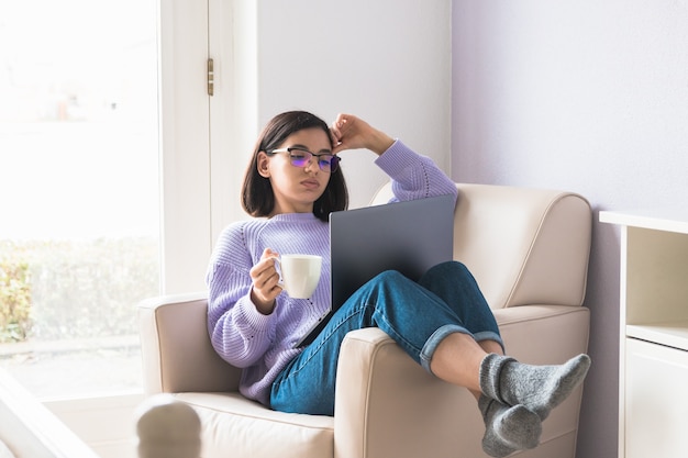 Verveeld om thuis te blijven. jonge professionele etniciteit vrouw of student in fauteuil in slaapkamer met een onlinevergadering en een kopje koffie te houden.