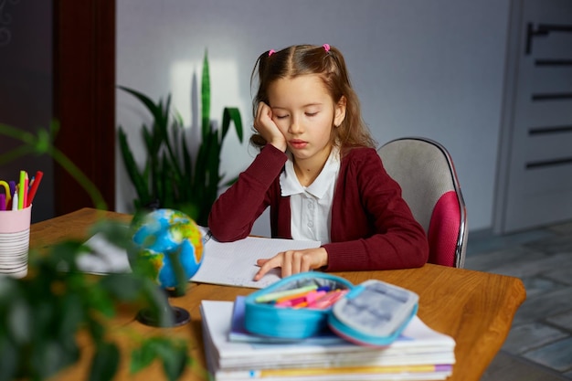 Verveeld schoolmeisje zit aan het bureau en doet huiswerk en leest thuisonderwijs