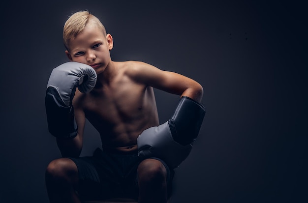 Verveelde jonge shirtless jongen die bokshandschoenen draagt die op een stoel zitten. geïsoleerd op een donkere achtergrond.