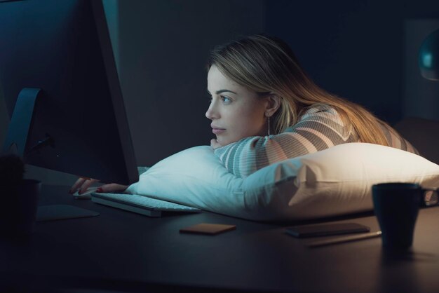 Verveelde vrouw die 's nachts overuren maakt, leunt op een kussen en typt op het toetsenbord van de computer