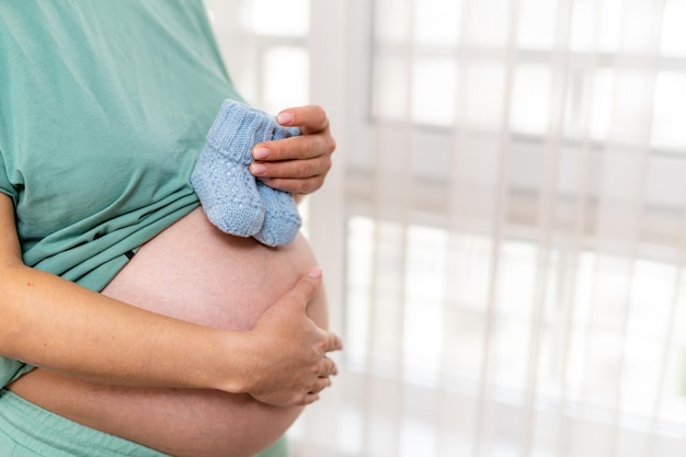 Verwacht een vrouw die bij het raam staat en babysokjes op haar buik houdt Dame die haar ene hand op de buik houdt en haar navel bedekt