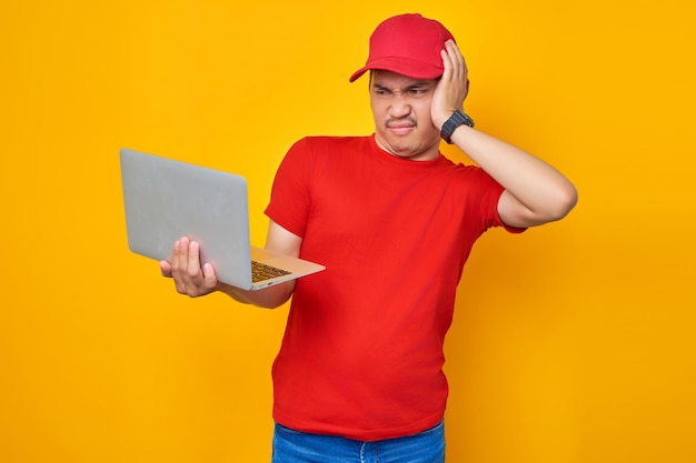 Verward jonge Aziatische man in rode dop tshirt uniforme werknemer werk als dealer koerier met behulp van laptop pc-computer op werk geïsoleerd op gele achtergrond Professional Delivery dienstverleningsconcept