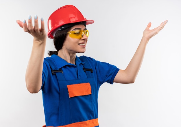 Verward kijkend naar de jonge bouwvrouw aan de zijkant in uniform met een bril die handen spreidt