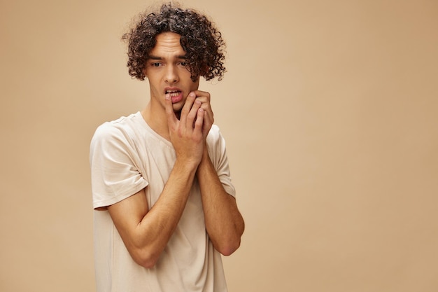 Verward ontzagwekkende gebruinde krullende man in basic tshirt raakt wangen poseren geïsoleerd op over beige pastel achtergrond Mode nieuwe collectie bieden mensen en emoties concept gratis plaats voor advertentie