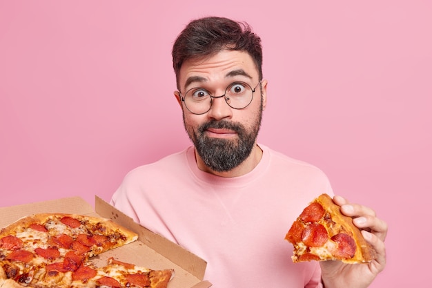 Verward verrast ongeschoren jonge man eet smakelijke pizza heeft fastfood-verslaving geniet van smakelijke snack in pizzeria kijkt verbaasd, poseert tegen roze muur. ongezond voedingsconcept