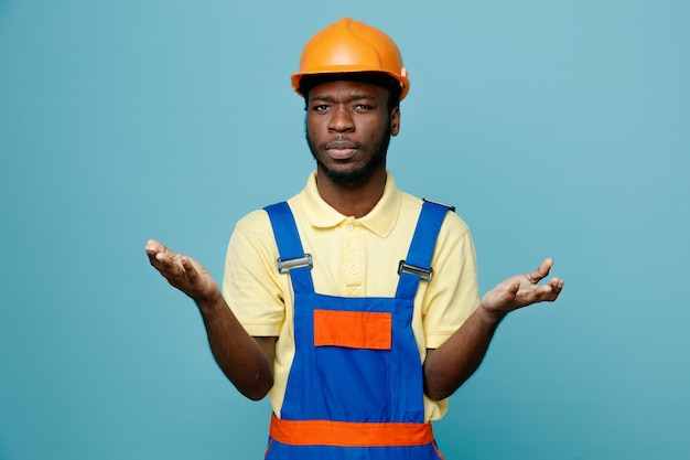 Verward verspreiden handen jonge Afro-Amerikaanse bouwer in uniform geïsoleerd op blauwe achtergrond