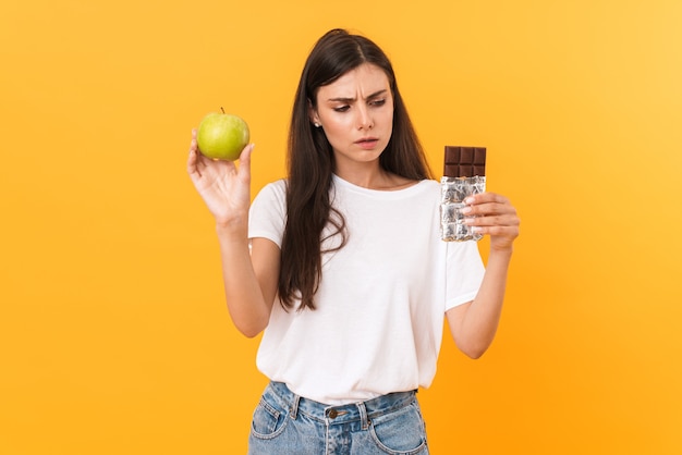 verwarde donkerbruine vrouw die basiskleren draagt die twijfel uitdrukken terwijl ze chocoladereep en groene appel vasthoudt die over gele muur wordt geïsoleerd