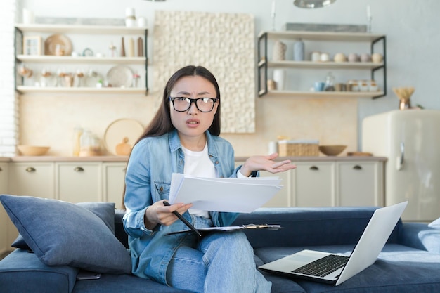 Verwarde en nerveuze jonge aziatische vrouw die thuis op de bank zit en met documenten en laptop werkt