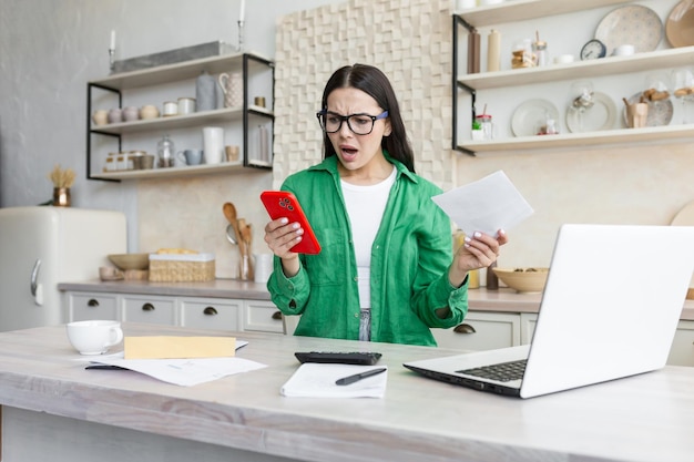 Verwarde en nerveuze jonge mooie vrouw met een bril die thuis in de keuken zit met een laptop