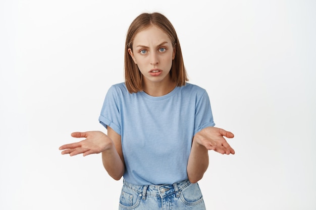 Verwarde en verbaasde blonde vrouw spreidde haar handen, haalde schouders op en fronste wenkbrauwen, vragend, niet begrijpend, staande tegen de witte muur.