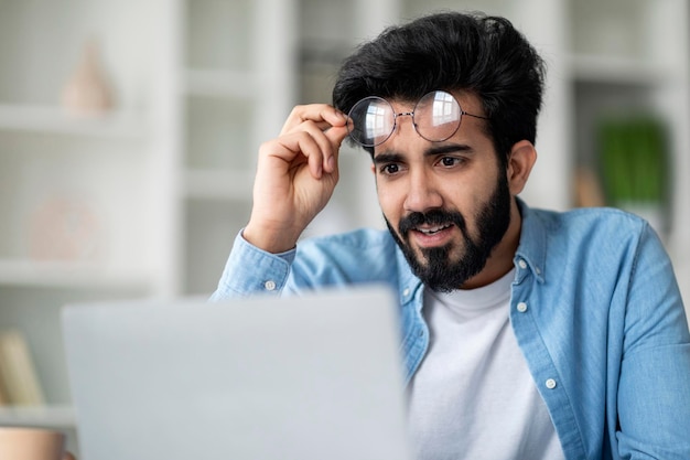 Verwarde Indiase mannelijke freelancer die een bril afdoet en naar het laptopscherm kijkt