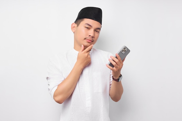Verwarde jonge Aziatische moslim man die mobiele telefoon vasthoudt terwijl hij de kin aanraakt en nadenkt over iets geïsoleerd op witte achtergrond Mensen religieus islamitisch levensstijlconcept