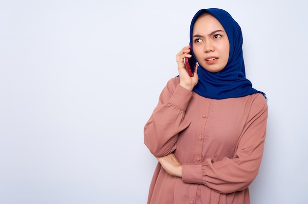 Verwarde jonge Aziatische moslimvrouw in roze shirt praten op mobiele telefoon geïsoleerd op witte achtergrond Mensen religieuze levensstijl concept